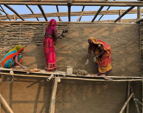 Drei Frauen in bunten Saris arbeiten auf einem Bambusgerüst an einer Lehmwand. Sie tragen Lehm auf die Wand auf, um eine Schule in Bahuarwa, Indien, zu bauen.