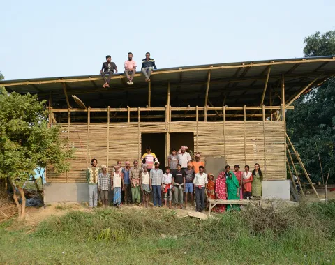 Eine Gruppe von Menschen steht vor einem im Bau befindlichen Gebäude aus Bambus in Bahuarwa, Indien. Drei Personen sitzen auf dem Dach. Das Bild zeigt den Fortschritt eines Schulbauprojekts, das von der Universität Liechtenstein unterstützt wird.