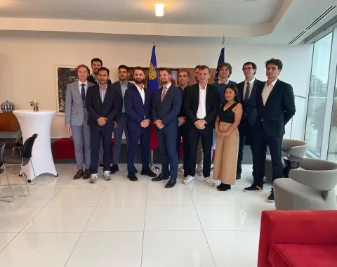 Gruppe von Studierenden und Begleitpersonen in Businesskleidung steht in einem modernen, hellen Raum mit Flaggen im Hintergrund.