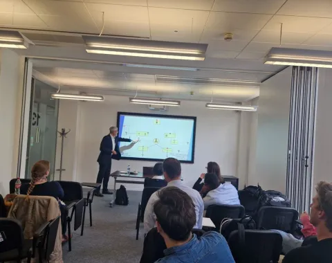 Ein Vortragender präsentiert vor einer Gruppe von Studierenden in einem modernen Seminarraum. Auf einem großen Bildschirm sind Diagramme zu sehen. Die Studierenden hören aufmerksam zu.