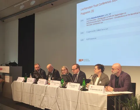 Ein Podium mit sechs Personen bei der Liechtenstein Trust Conference 2024. Sie sitzen an einem Tisch mit Mikrofonen und Namensschildern. Im Hintergrund ist eine Leinwand mit dem Konferenzprogramm zu sehen.