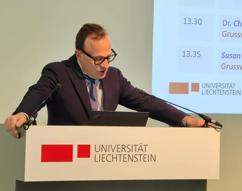 Ein Redner steht am Podium der Universität Liechtenstein und hält eine Präsentation. Im Hintergrund ist eine Projektionsfläche mit einem Zeitplan zu sehen.