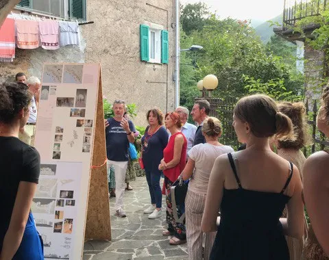 Eine Gruppe von Menschen betrachtet eine Ausstellung im Freien in einem historischen Dorf. Im Vordergrund sind Informationstafeln zu sehen, im Hintergrund eine steinerne Hausfassade und grüne Hügel.