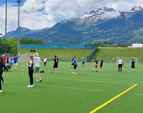 Eine Gruppe von Menschen spielt Völkerball auf einem grünen Sportplatz vor einer beeindruckenden Bergkulisse. Der Himmel ist teils bewölkt. Die Szene zeigt sportliche Aktivität und Gemeinschaftsgefühl bei sonnigem Wetter.