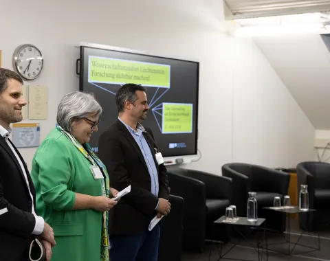 Drei Personen stehen nebeneinander in einem Konferenzraum. Im Hintergrund ist ein Bildschirm mit der Aufschrift "Wissenschaftsverbund Liechtenstein" zu sehen. Links im Bild ist ein Rednerpult, rechts sind leere Stühle und Tische. Die Personen tragen Namensschilder und lächeln.