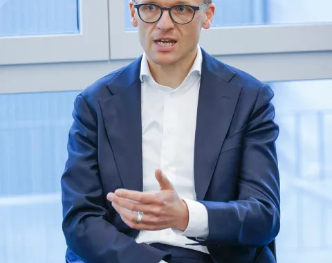 Ein Mann in einem Anzug spricht gestikulierend während einer Podiumsdiskussion in einem modernen Raum mit großen Fenstern.