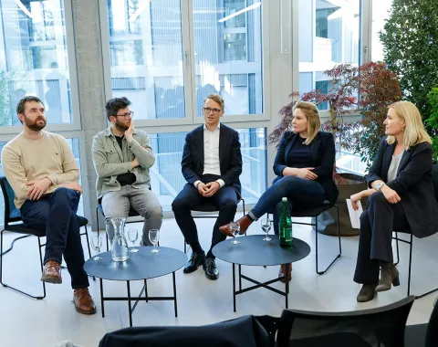 Podiumsdiskussion an der Universität Liechtenstein: Fünf Personen sitzen in einem modernen Raum mit großen Fenstern und Pflanzen, diskutieren über den Wohnungsmarkt. Vor ihnen stehen Tische mit Wassergläsern und einer Flasche.