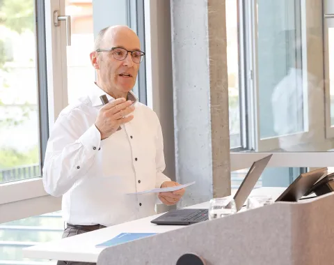 Ein Mann hält einen Vortrag in einem modernen Raum mit großen Fenstern. Er steht an einem Rednerpult mit Laptop und Notizen.