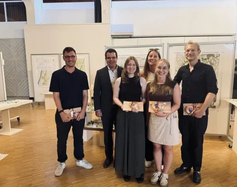 Gruppe von Studierenden und Dozenten der Universität Liechtenstein präsentiert Architekturprojekte im Rahmen eines Advanced Studios. Im Hintergrund sind Plakate und Modelle zu sehen, die ortsbauliche Strategien für die Gemeinde Ruggell zeigen.