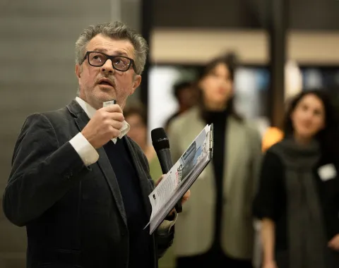 Ein Mann mit einer schwarzen Brille im Anzug spricht in ein Mikrofon