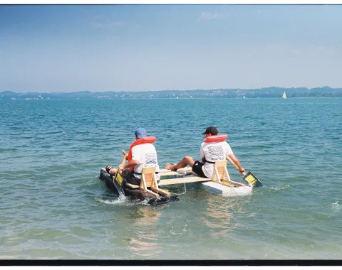 Zwei Menschen mit Schwimmweste in einem selbstgebauten Boot. 