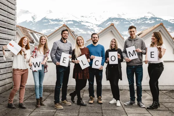Eine Gruppe junger Menschen steht nebeneinander auf einer Terrasse vor schneebedeckten Bergen und hält Buchstaben-Schilder hoch, die zusammen das Wort "WELCOME" ergeben. Sie lächeln und wirken freundlich, in legerer Kleidung. Der Hintergrund zeigt schneebedeckte Berge und einige Gebäude mit Giebeldächern, was auf eine winterliche oder alpine Umgebung hindeutet.