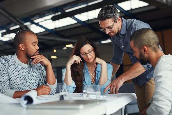 Eine Gruppe von vier Menschen, die um einen Tisch in einem modernen Büro sitzen und gemeinsam an einem Projekt arbeiten. Zwei Männer, eine Frau und ein weiterer Mann, der auf das Dokument zeigt, sind in eine Diskussion vertieft. Vor ihnen liegen Pläne und Dokumente, auf dem Tisch stehen Gläser mit Wasser. Die Atmosphäre wirkt konzentriert und kollaborativ.