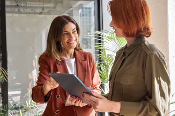 Zwei Frauen stehen in einem modernen Bürogebäude und unterhalten sich. Beide sind formell gekleidet und wirken konzentriert. Im Hintergrund sind große Fenster zu sehen, die viel natürliches Licht in den Raum lassen, sowie Büromöbel und Pflanzen, die eine professionelle Atmosphäre schaffen.