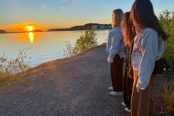 Drei Studentinnen der Universität Liechtenstein beobachten gemeinsam den Sonnenuntergang an einem ruhigen Seeufer.