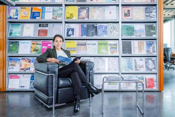 Frau sitzt entspannt auf einem schwarzen Ledersessel in der Bibliothek der Universität Liechtenstein und liest in einer Fachzeitschrift, im Hintergrund ein Regal mit bunten Magazincovern.