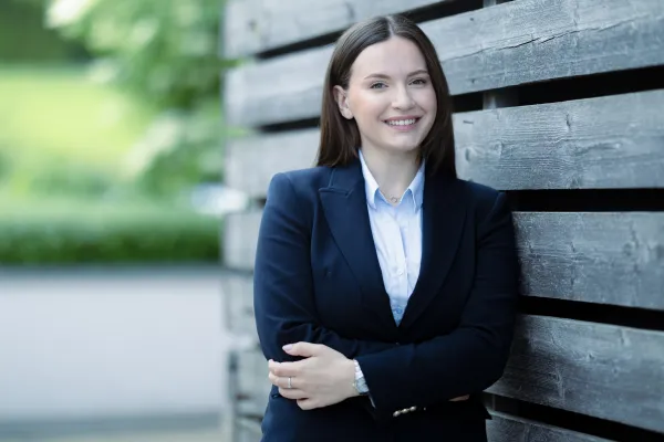Eine weibliche Studierende im Masterstudiengang Innovative Finance im dunkelblauen Blazer steht mit verschränkten Armen vor einer Holzfassade und lächelt freundlich.