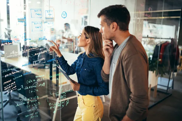 Ein Mann und eine Frau betrachten gemeinsam eine große Glaswand, die mit Notizen und Diagrammen beschrieben ist. Beide wirken nachdenklich und konzentriert, während sie an einer komplexen Aufgabe arbeiten.