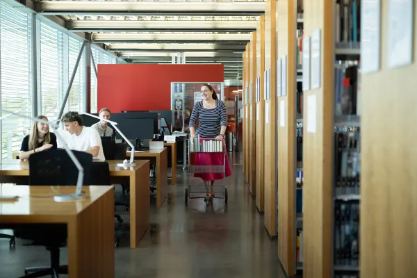 Studierende arbeiten und unterhalten sich in der Bibliothek, während die Bibliotheksmitarbeiterin mit Bücherwagen vorbeigeht.
