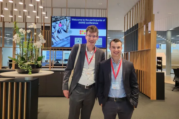 Zwei Männer in Anzügen mit roten Namensschildern stehen lächelnd in einem modernen Konferenzraum. Im Hintergrund ist ein Bildschirm mit der Aufschrift „Welcome to the participants of AAWE conference“ zu sehen. Links steht eine dekorative Pflanze auf einem Tisch.