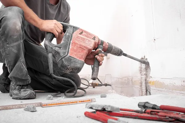 Ein Bauarbeiter kniet auf dem Boden und benutzt einen roten Kombi-Hammer, um eine Wand zu bearbeiten. Im Vordergrund liegen Werkzeuge und Bauutensilien.