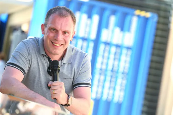 Andreas Kräätli lächelnd in einem grauen Polohemd hält ein Mikrofon. Im Hintergrund sind verschwommene blaue Strukturen zu sehen.