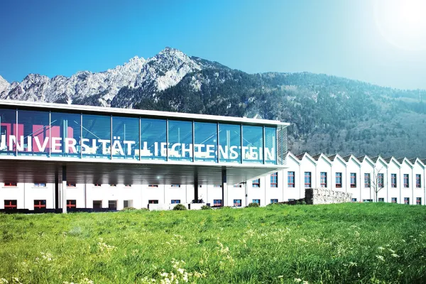 Außenansicht der Universität Liechtenstein mit modernem Glasgebäude vor malerischer Bergkulisse, umgeben von grüner Wiese.