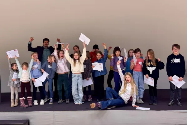 Eine Gruppe von Kindern und einem Erwachsenen steht auf einer Bühne und hält stolz Zertifikate hoch. Die Kinder wirken fröhlich und begeistert. Sie nehmen an einem Fundraising-Workshop an der Universität Liechtenstein teil.
