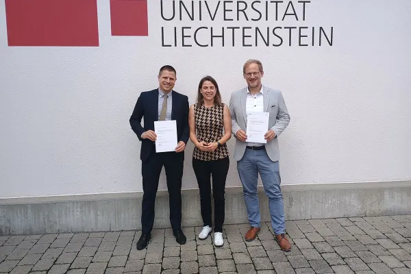 Drei Personen stehen vor einer Wand mit dem Logo der Universität Liechtenstein. Zwei von ihnen halten Zertifikate in den Händen.