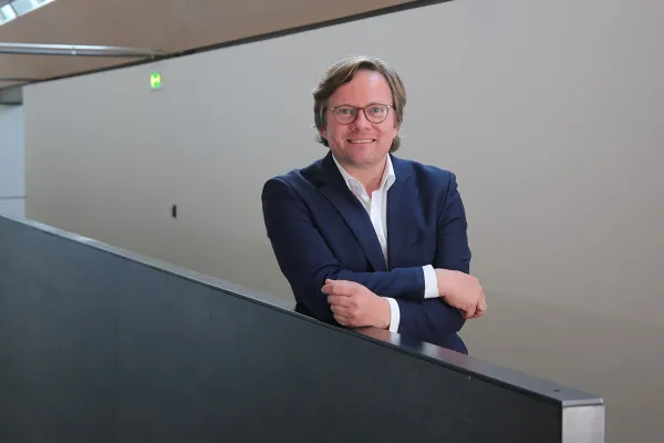 Ein Mann in einem blauen Anzug lehnt lächelnd an einem Geländer in einem modernen Gebäude.