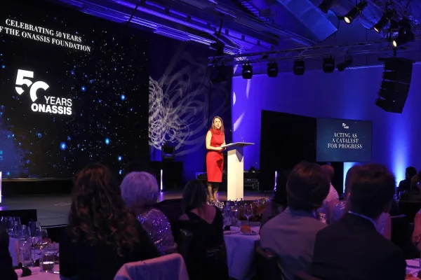 Eine Rednerin in rotem Kleid steht an einem Podium auf einer Bühne, umgeben von Publikum. Im Hintergrund ist eine Leinwand mit der Aufschrift "Celebrating 50 Years of the Onassis Foundation" zu sehen. Die Veranstaltung findet in einem festlich beleuchteten Raum statt.