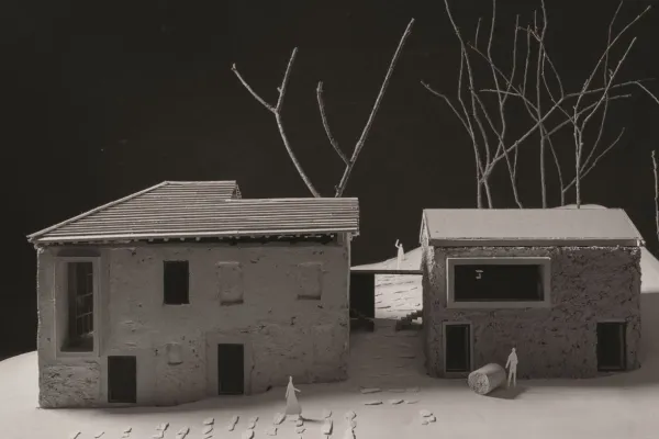 Modell eines architektonischen Projekts mit zwei Gebäuden, umgeben von kahlen Bäumen. Das linke Gebäude hat ein geneigtes Dach, während das rechte ein Flachdach aufweist. Kleine Figuren und Fußspuren sind auf dem Boden sichtbar, was eine menschliche Präsenz andeutet.