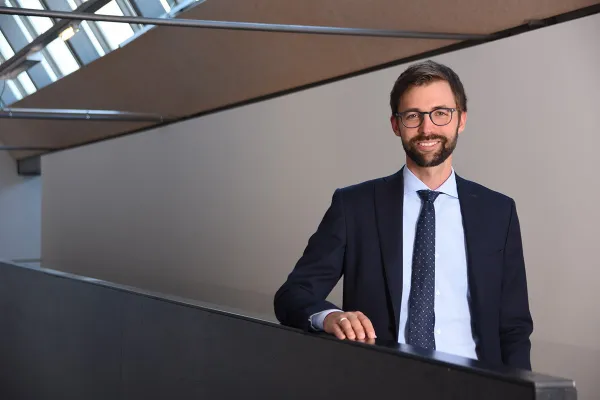 Ein Mann in Anzug und Krawatte steht lächelnd in einem modernen Gebäude mit schrägen Decken und großen Fenstern.