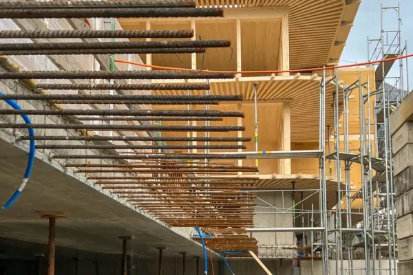 Baustelle eines modernen Gebäudes mit sichtbaren Stahlträgern und Holzverkleidung, umgeben von Gerüsten, unter bewölktem Himmel.