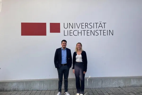 Zwei Personen stehen vor einer Wand mit dem Logo der Universität Liechtenstein. Sie lächeln in die Kamera.