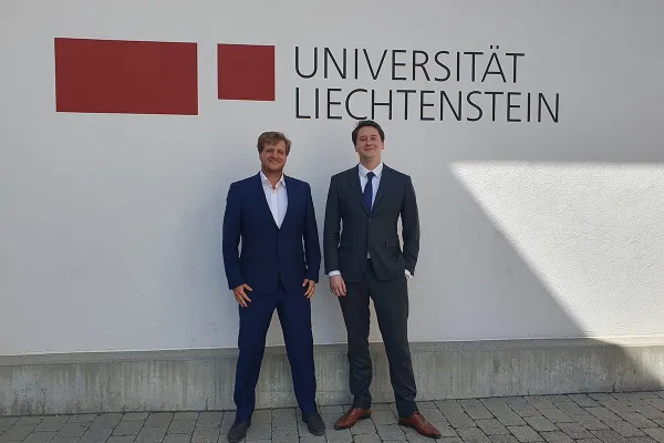Zwei Personen in Anzügen stehen vor einer Wand mit dem Logo der Universität Liechtenstein.