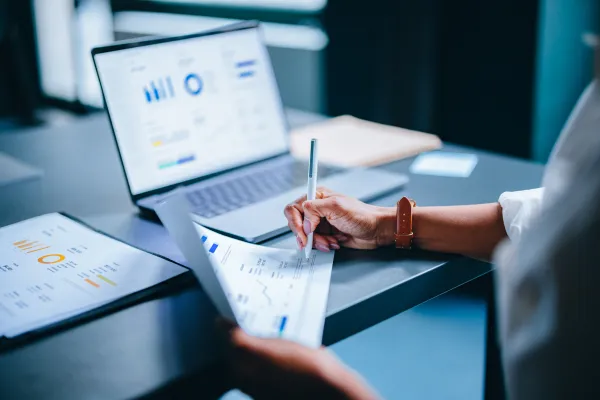 Hand schreibt Notizen zu Finanzstatistiken vor geöffnetem Laptop mit Diagrammen und Tabellen.