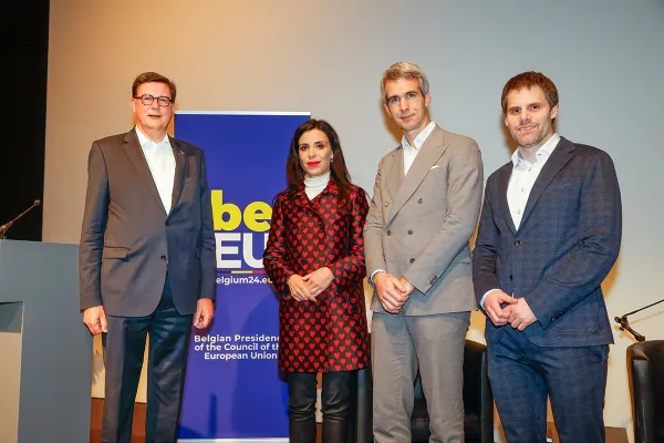 Vier Personen stehen nebeneinander vor einem Banner mit der Aufschrift "Belgian Presidency of the Council of the European Union". Sie tragen formelle Kleidung und blicken in die Kamera.