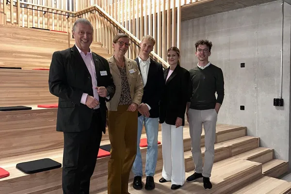 Fünf Personen stehen lächelnd in einer modernen Aula mit Holzstufen und vertikalen Holzlamellen im Hintergrund. Sie tragen formelle Kleidung und posieren für ein Gruppenfoto.