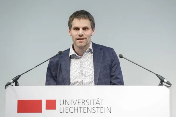 Rektor Christian Frommelt hält eine Rede am Rednerpult vor einem sitzenden Publikum in einem Konferenzraum. Im Hintergrund ist ein Poster sichtbar.