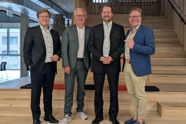 Vier Männer in formeller Kleidung stehen lächelnd auf einer Holztreppe in einem modernen Gebäude.