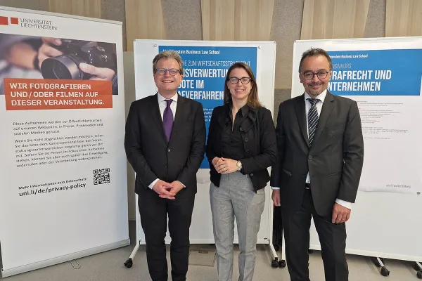 Drei Personen stehen lächelnd vor Informationsplakaten der Universität Liechtenstein. Die Plakate thematisieren Wirtschaftsstrafrecht und Beweisverwertungsverbote. Die Personen tragen formelle Kleidung.