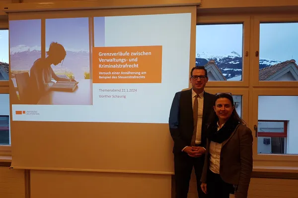 Ein Mann und eine Frau stehen vor einer Präsentationsleinwand mit dem Titel "Grenzverläufe zwischen Verwaltungs- und Kriminalstrafrecht" im Seminarraum der Universität Liechtenstein. Im Hintergrund sind schneebedeckte Berge durch die Fenster sichtbar.